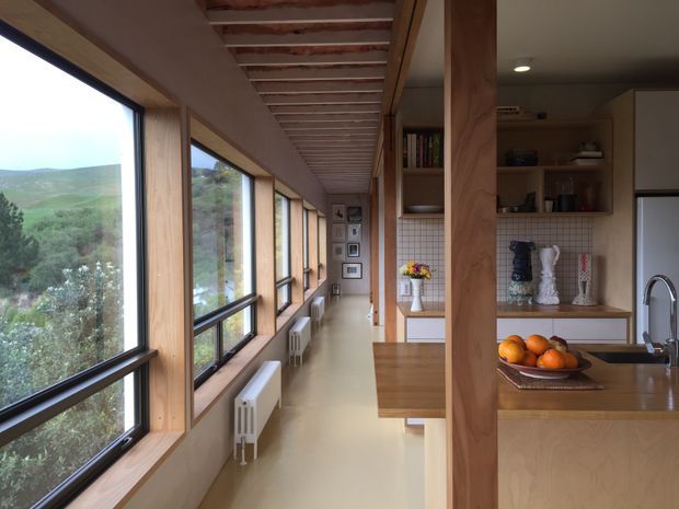 A long, narrow open-plan kitchen and circulation space with extensive timber-framed glazing along one side, revealing a green hilly landscape. The kitchen has light timber cabinetry and benchtops, a stainless-steel sink and tap, open shelving, a tiled splashback, and a timber post dividing the view. Exposed ceiling battens run along the length of the room, with radiators beneath the windows and a small dining or work surface in the foreground.