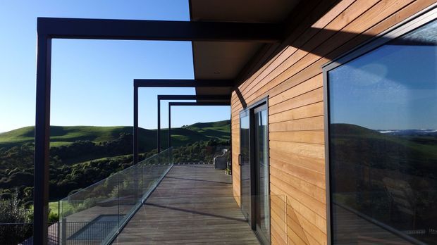 Exterior view of a contemporary timber-clad building with a long elevated deck or balcony, frameless glass balustrade, dark metal rectangular portal frames, and large glazed openings. The structure overlooks rolling green hills in bright natural light.