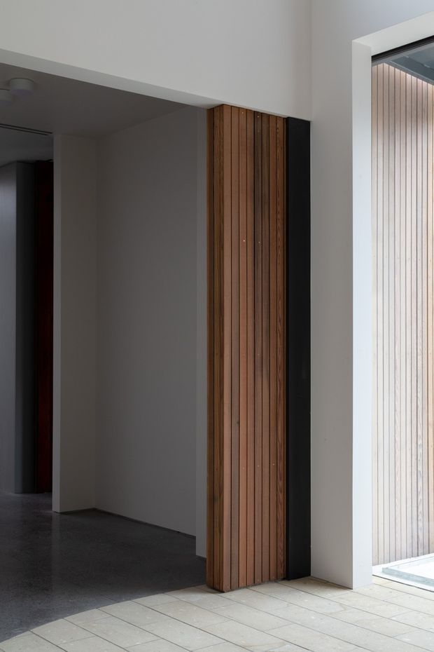 Minimalist architectural entry or circulation space with white plaster walls, a dark polished concrete floor, and a prominent vertical timber slat feature panel framed in black. A large glazed opening on the right reveals a matching light timber slatted exterior screen and brings in strong natural light. The image emphasizes clean geometric lines, contrasting timber and monochrome surfaces, and a restrained contemporary material palette.