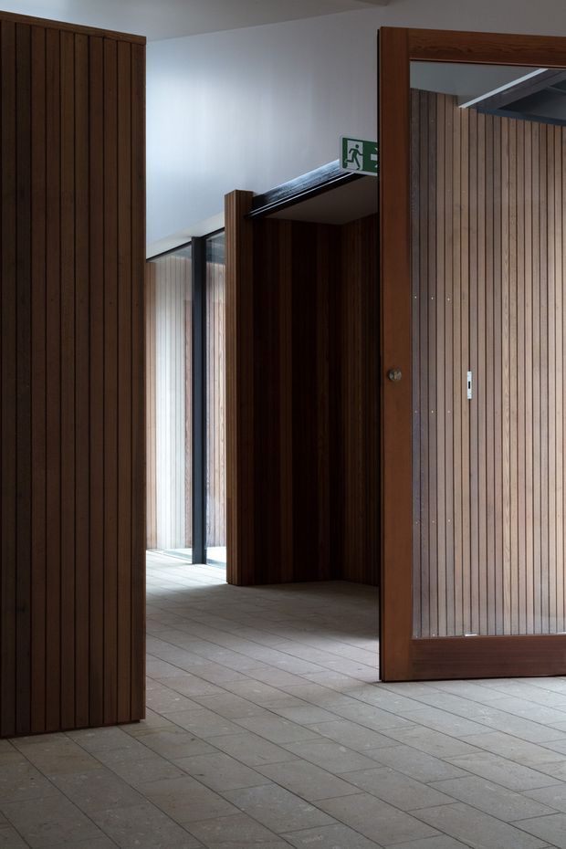 A quiet institutional or commercial interior corridor with tall dark timber slatted wall panels and a large timber-framed glazed door. Pale square floor tiles, white walls, black-framed glazing, and an illuminated green emergency exit sign create a clean, understated architectural setting.