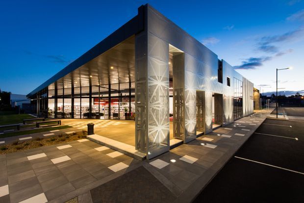 Exterior dusk view of a contemporary commercial building with a long, angular form, extensive floor-to-ceiling glazing, metallic panel cladding, and illuminated patterned feature screens at the entrance. A paved pedestrian forecourt with benches and planted strips fronts the glazed interior, while marked parking and street lighting extend along the side.