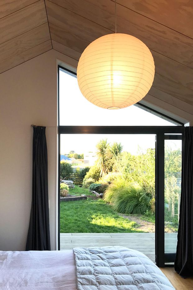 A bright bedroom viewed toward a large black-framed glazed opening and adjoining glass door leading to a timber deck and landscaped garden. The room has a warm timber-lined pitched ceiling, pale walls, timber flooring, dark curtains, and a large glowing spherical pendant light. The lower foreground shows part of a bed with pale bedding and a grey quilt. The glazing provides strong indoor-outdoor connection and abundant natural light.