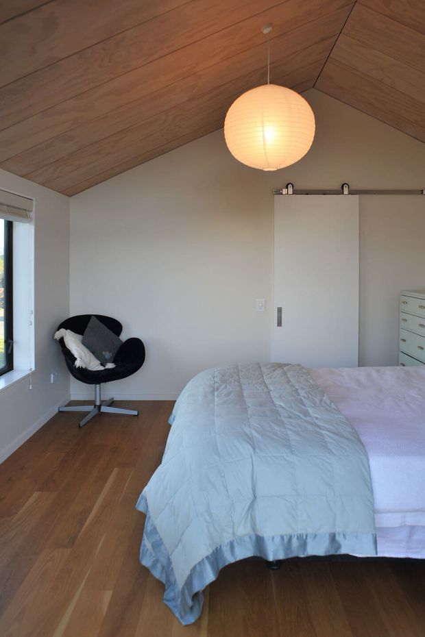 A compact, calm bedroom with a pitched ceiling clad in warm timber boards, pale walls, timber flooring, and a large bed dressed in soft pale blue and white bedding. A black lounge chair sits beside the window, while a simple white sliding barn-style door and a light chest of drawers provide functional storage. A round paper pendant light hangs centrally from the ceiling.