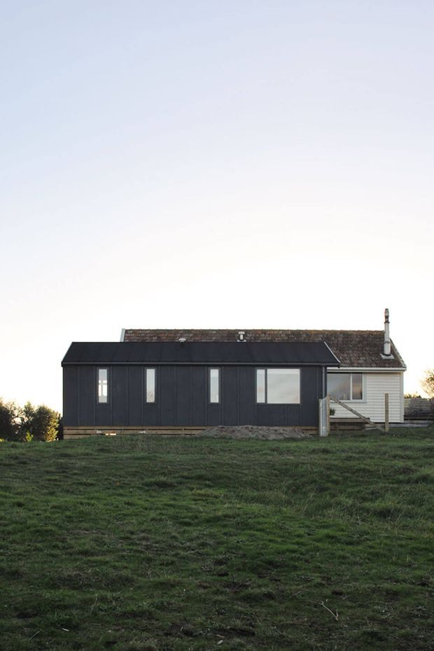 A small single-storey rural house viewed from across a gently sloping grassy field. The building combines a long dark charcoal-clad extension with narrow vertical windows and a pitched roof, attached to a lighter weatherboard section with a tiled roof, chimney, and small exterior steps. The scene has a simple, utilitarian character and subdued natural lighting.