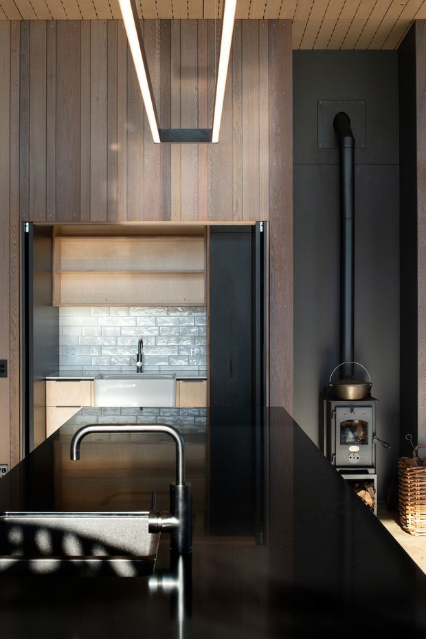 A compact, dark-toned kitchen with a black island and integrated stainless-steel sink and tap in the foreground. The rear kitchenette features pale timber cabinetry, open shelving, a white farmhouse-style sink, and a glossy white subway-tile splashback. Vertical dark timber wall panelling and a timber slatted ceiling frame the space, while a black wood-burning stove and flue occupy the right side. A linear pendant light hangs overhead, creating a warm, modern-rustic atmosphere.
