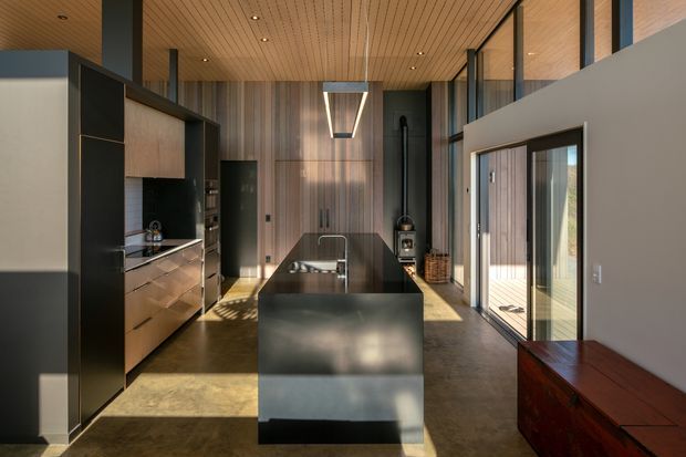 A sophisticated open-plan kitchen with a large dark island, integrated sink and tap, dark cabinetry, warm timber wall and ceiling lining, and polished concrete flooring. Full-height glazing and sliding doors bring in abundant natural light, while a contemporary linear pendant hangs above the island. The space has a tall, airy volume and refined, minimalist detailing.