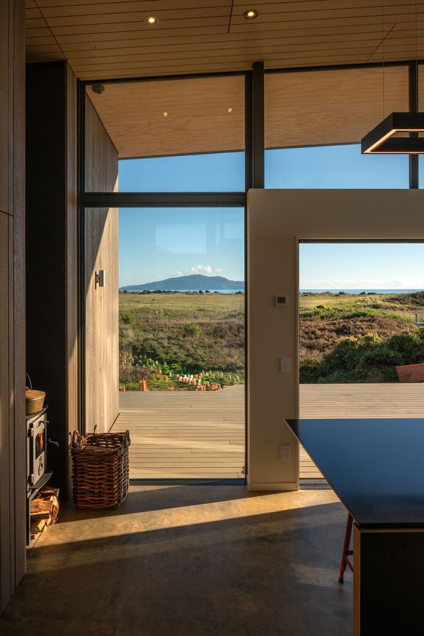 A warm, modern interior opening onto a timber deck and expansive coastal landscape. The room features full-height dark-framed glazing and doors, a timber-lined ceiling, polished concrete flooring, recessed lighting, and a dark kitchen or work counter at right. A compact wood-burning stove and woven basket sit along the left wall. The view includes open coastal vegetation, water, and a distant mountainous island or headland.