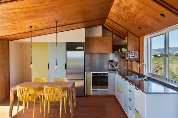 A compact open-plan kitchen and dining area with extensive warm timber lining across the ceiling and upper walls. The kitchen has pale blue-grey and muted green fixed cabinetry, stainless-steel appliances and splashback, dark stone-look benchtops, and a large window overlooking a rural landscape. A timber dining table with bright yellow chairs sits beneath simple pendant lights.