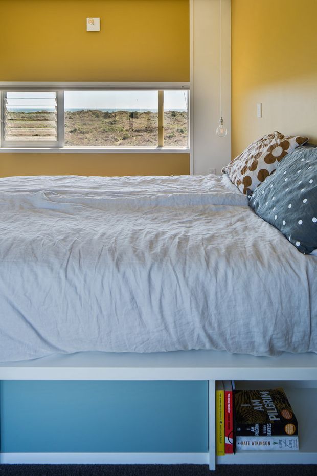 A compact, bright bedroom with a low platform bed occupying most of the frame. The bed is dressed in a light blue-grey rumpled duvet with patterned pillows, set against mustard yellow and pale neutral walls. A wide horizontal window provides expansive coastal landscape views, while open shelving beneath the bed contains books and adds a practical built-in storage feature. The room has a playful, casual contemporary feel with strong natural light and a blue accent panel below the bed.
