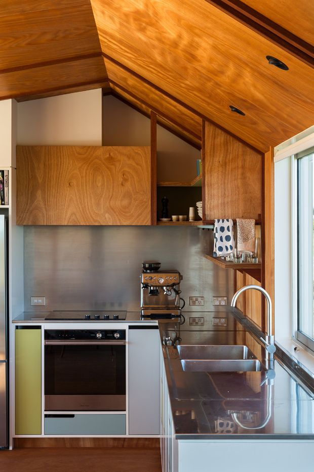 A compact, highly detailed kitchen with custom warm timber cabinetry extending across the walls and ceiling, stainless-steel splashback, dark reflective benchtops, integrated oven and cooktop, espresso machine, and a deep double-bowl sink beneath a large window. The cabinetry has strong grain patterns and black pulls, while a muted lime-green cabinet panel adds a small accent.