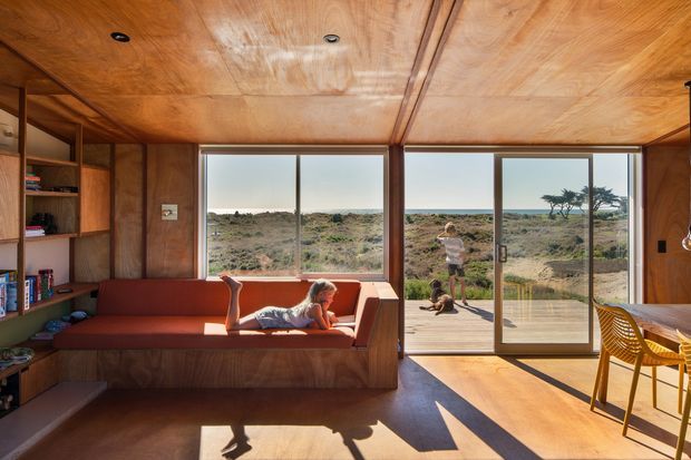 A warm, compact timber-lined living room opening onto a timber deck and coastal landscape. The room features built-in open shelving, a large built-in window seat with a rust-red cushion, expansive sliding glass doors, a timber ceiling with recessed downlights, and a small dining area with woven chairs. Strong sunlight creates long shadows across the timber-toned floor, giving the space a relaxed, indoor-outdoor character.
