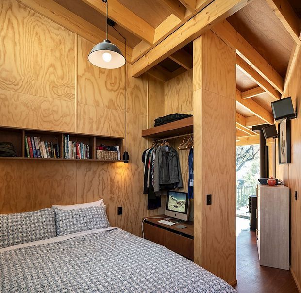Compact bedroom clad in warm plywood, with a patterned bed, built-in open shelving filled with books, an integrated wardrobe and desk nook, exposed timber beams, and a passage leading toward a bright glazed exterior opening. A simple black pendant light and wall-mounted fixtures complement the practical timber interior.