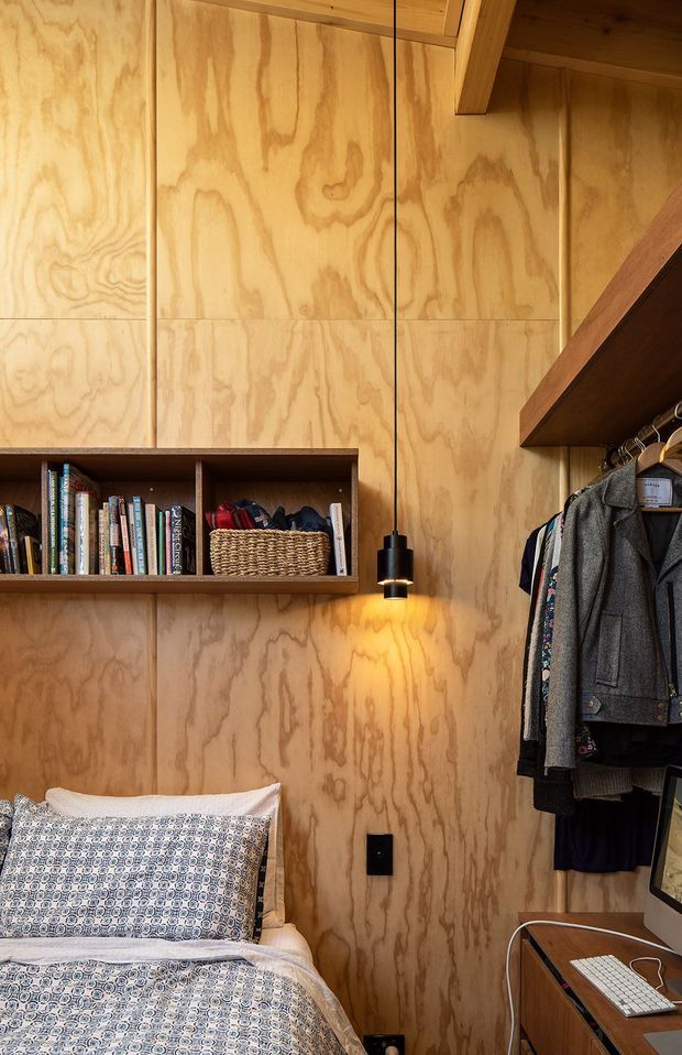 Compact bedroom with full-height plywood wall panelling, exposed timber ceiling structure, built-in open bookshelf, hanging pendant light, wall-mounted clothing rail, and a bed with patterned bedding. A small desk and computer are visible at the lower right. The warm timber-lined interior has a practical, built-in storage-focused design.