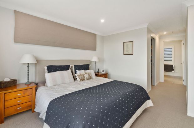 Bright, comfortably furnished bedroom with a large bed dressed in white bedding and a dark navy patterned throw. The room has light neutral walls, beige carpet, white trim, warm timber bedside drawers, bedside lamps, and a hallway leading to additional rooms. A roller blind covers the main window, with a second shuttered window visible down the hall.