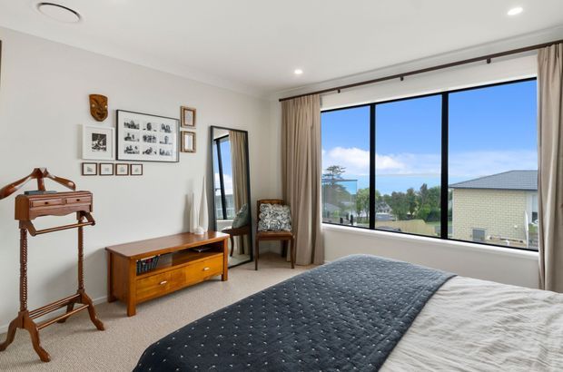 Bright, neatly styled bedroom with a large black-framed picture window overlooking a suburban landscape. The room features a bed with a dark quilt, beige carpet and curtains, a mid-century timber television console, vintage valet stand, framed wall art, and a full-length mirror. Recessed ceiling lights and a simple neutral palette create a calm, comfortable atmosphere.