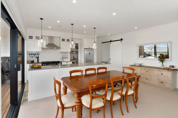 Bright open-plan kitchen and dining room with white cabinetry, a large island, pendant lighting, and a traditional polished timber dining suite. A sliding glass door opens to the exterior, while a timber sideboard and barn-style door hardware add character.