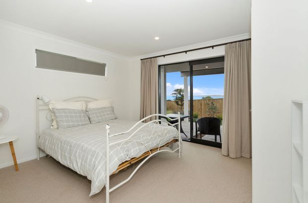 Bright, compact bedroom with a white metal bed frame, light grey checked bedding, pale carpet, white walls and traditional cornice detailing. Full-height curtains frame black-framed glazed doors opening to a small outdoor terrace or balcony, with a distant coastal landscape visible beyond.