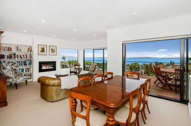 Bright open-plan dining and living area with a polished traditional timber dining table and chairs in the foreground, a leather sofa and patterned armchair near a built-in fireplace, and extensive glazing opening to a timber deck with a coastal ocean view. The room has white walls, recessed lighting, carpeted flooring, and built-in bookshelves.