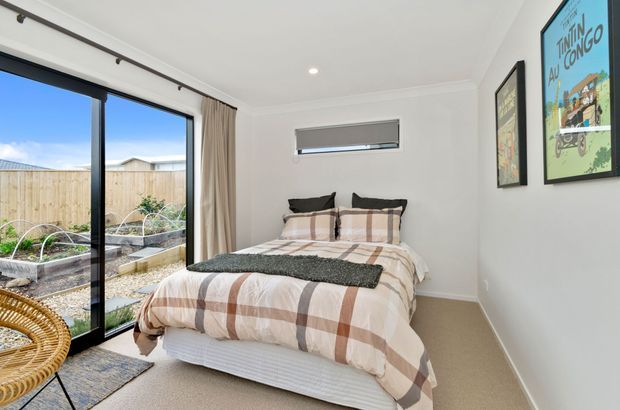 Bright, compact modern bedroom with a neatly made bed, neutral plaid bedding, beige carpet, white walls and ceiling, and framed artwork. Full-height black-framed sliding glazing opens onto a small landscaped garden with raised planting beds, while a narrow horizontal window provides additional daylight. A woven chair and small rug add natural texture.