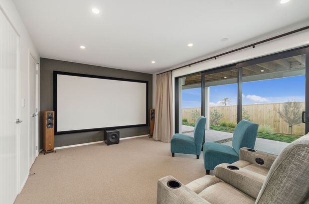 A dedicated home cinema/media room with a large wall-mounted projection screen, floor-standing speakers, subwoofer, and multiple upholstered cinema recliners. Full-height glazing opens the room to a small landscaped outdoor area, while beige carpet, dark grey feature wall, white ceiling, and recessed lighting create a clean contemporary setting.
