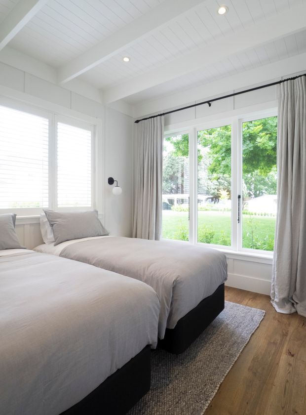 Bright, calm bedroom featuring two twin beds with soft light-grey bedding and black upholstered bases. The room has white painted walls and a white slatted vaulted ceiling with exposed beams, plantation shutters, floor-to-ceiling glazed doors, full-height neutral curtains, warm timber flooring, and a grey textured rug. Green garden views are visible through the glazing.