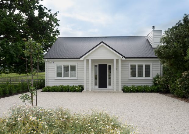 A charming single-storey cottage-style home viewed from the front, with white horizontal weatherboard cladding, a dark grey pitched metal roof, a central gabled entry porch, white-framed sash-style windows, and a pale gravel driveway bordered by landscaped planting.