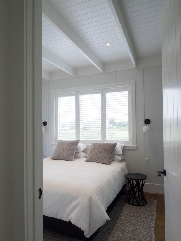 Bright, compact bedroom viewed through an open doorway, featuring a white upholstered or neatly made bed, textured neutral cushions, plantation-shuttered windows, white vertical wall panelling, and a white beadboard ceiling with exposed beams. Black wall sconces and a small black side table provide contrast, while timber flooring and a neutral rug add warmth.