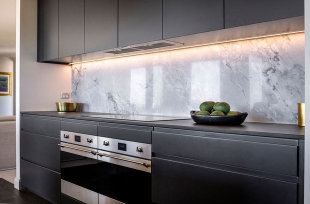 A sleek contemporary kitchen featuring matte dark-grey full-height cabinetry, integrated appliances, a pale marble-look splashback with prominent veining, and warm under-cabinet lighting. The long, streamlined benchtop includes a decorative bowl and minimal accessories.
