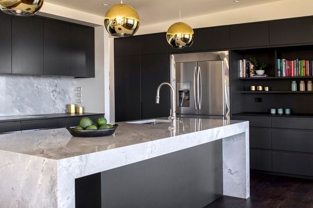 Contemporary kitchen featuring extensive matte black cabinetry, a large white and grey veined stone island with waterfall ends, stainless-steel refrigerator, integrated shelving, and three spherical brass pendant lights. Dark timber flooring and a pale ceiling create a high-contrast, refined interior.