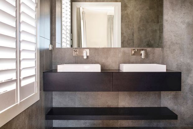 Contemporary double-vanity bathroom with two white vessel basins, wall-mounted dark timber cabinetry, a matching open shelf, brushed metal wall-mounted tapware, large grey stone-look wall tiles, and a white-framed mirror. Plantation shutters provide filtered daylight on the left.