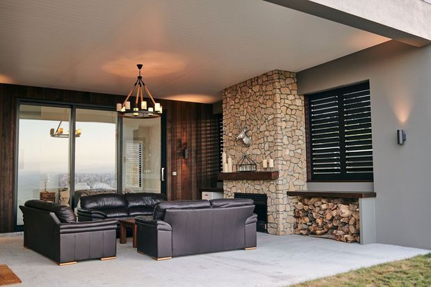 Covered outdoor living area with dark leather seating arranged around a stone fireplace and hearth. The space has a pale concrete floor, a timber-lined wall, exposed natural stonework, dark louvred windows, a statement pendant light, and expansive glazing overlooking a distant cityscape.