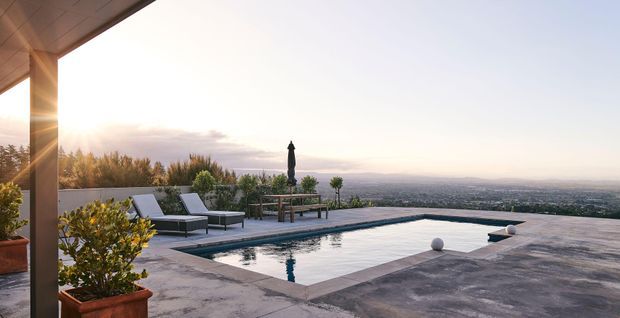 A rooftop or elevated outdoor terrace with a rectangular swimming pool, lounge chairs, dining table, potted planting, and broad distant landscape views. The space has a warm sunset atmosphere and a restrained contemporary finish with large-format grey paving.