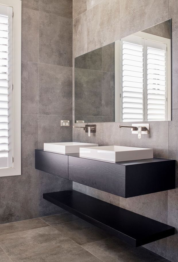 Contemporary bathroom with large grey stone-look wall and floor tiles, a wall-mounted dark timber vanity with two white vessel basins, wall-mounted chrome taps, a large frameless mirror, and white plantation shutters.