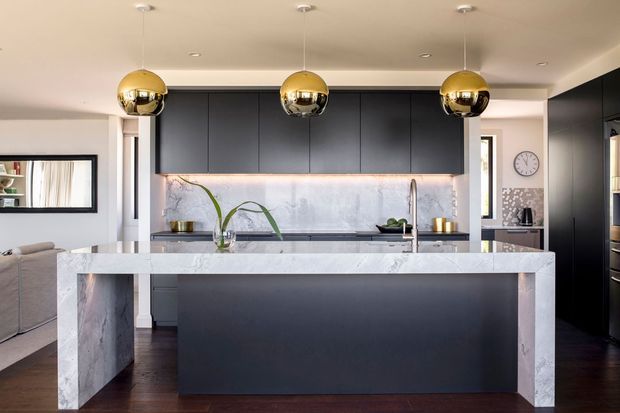 Contemporary kitchen featuring a large marble-clad island with dark cabinetry, a pale stone splashback, integrated appliances, and three reflective brass pendant lights. A living area is visible to the left and a dining or hallway area beyond the kitchen.