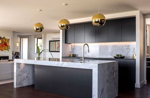 Contemporary open-plan kitchen with a large waterfall-edge island in dark cabinetry and veined grey stone, matching full-height splashback, and sleek black built-in cabinetry. Three spherical brass pendant lights hang above the island. A living area is visible beyond the kitchen, with dark timber flooring, neutral walls, and restrained modern furnishings.