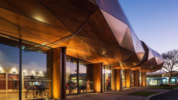 Exterior dusk view of a contemporary commercial building with a dramatic faceted, angular metal canopy and warm timber soffit supported by structural columns. Floor-to-ceiling glazing reveals retail or public interior spaces, while illuminated entrances line the paved frontage.