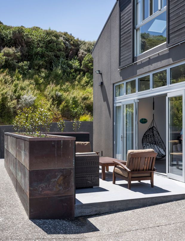 A sunny exterior patio beside a contemporary multi-level home, with dark horizontal cladding, rendered walls, extensive glazed doors and windows, and a sheltered seating area. The patio includes a large rusted steel raised planter, woven outdoor seating, a timber armchair and side table. Dense native planting rises behind the terrace, and the interior is visible through the open sliding doors.