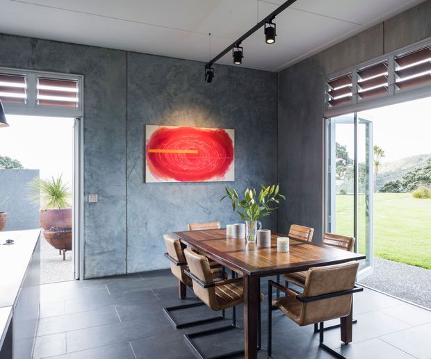 A contemporary dining room with a dark textured concrete-look feature wall, large tiled floor, timber dining table and leather-upholstered chairs. Full-height glazed openings with external louvres connect the room to a bright lawn and garden, while track lighting and vibrant abstract artwork add visual contrast.
