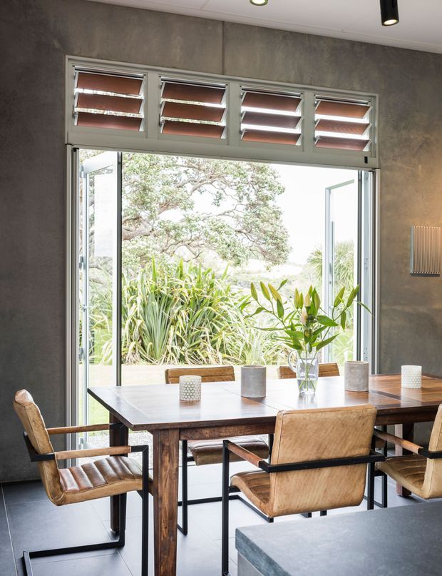 A contemporary dining area with a dark timber dining table and leather-upholstered armchairs, positioned beside large open bifold-style glazing. Above the opening are rows of louvred awning windows. The room has textured dark grey walls, a light ceiling with recessed and track lighting, a dark tiled floor, and a stone-topped counter in the foreground. Lush planting is visible outside.