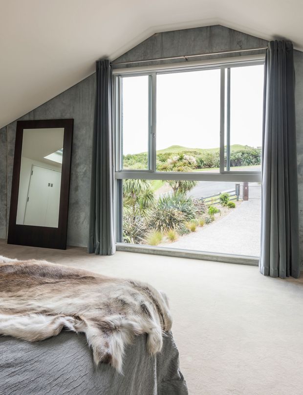 A contemporary bedroom with a dramatic angled ceiling and large floor-to-ceiling window overlooking landscaped grounds. The room features grey concrete-look wall panelling, pale flooring, full-length charcoal curtains, a dark-framed leaning mirror, and a bed with a neutral grey cover and textured fur throw.