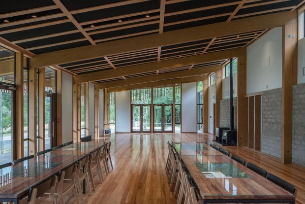 A long, expansive contemporary interior with a central timber floor and exposed timber structural framing. Glass-topped dining or meeting tables with numerous chairs line both sides, while floor-to-ceiling glazing opens views to greenery outside. The ceiling features a dramatic exposed timber grid with recessed lighting, and the walls combine light plasterboard with sections of grey masonry. The space is bright, warm, and strongly connected to the outdoors.