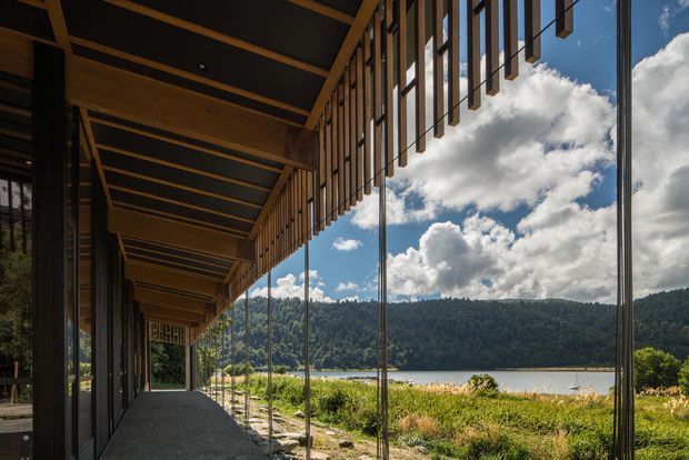 A covered architectural walkway or terrace runs along a glazed building edge, with exposed timber roof framing and slender vertical timber sun-shading battens. Floor-to-ceiling glazing opens views toward a lake, wooded hills and a bright sky. The structure has a dark, contemporary material palette with warm timber detailing and a strong indoor-outdoor connection.