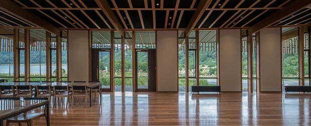 A spacious, sophisticated open-plan interior with extensive floor-to-ceiling glazing, a polished timber floor, and a dramatic exposed timber ceiling grid. Dining tables and chairs occupy the left side, while the room opens toward a lush landscaped exterior visible through the windows. The warm timber structure and reflective floor create a refined, airy atmosphere.
