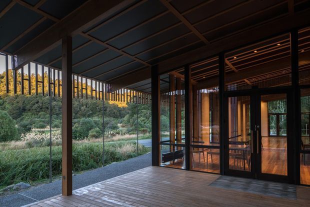 A covered timber deck or veranda adjoining a modern glazed building. Floor-to-ceiling glass doors and windows reveal a warmly lit interior with timber flooring, while dark timber posts, beams and a gridded ceiling structure frame views toward landscaped vegetation and a hillside. The space has a strong indoor-outdoor connection and a calm, natural atmosphere.