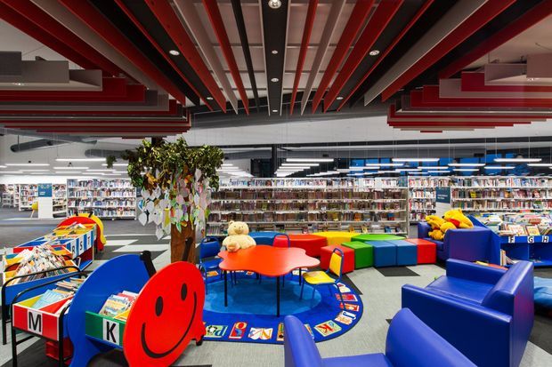A brightly coloured children’s reading and activity area inside a large library or educational commercial space. The foreground contains blue lounge chairs, red and orange activity tables, colourful modular seating, children’s books, alphabet-themed floor graphics, and a playful tree-like display. Long rows of bookshelves extend across the background. The ceiling features exposed red, black, and grey linear beams with integrated recessed lighting, creating a bold contemporary interior.