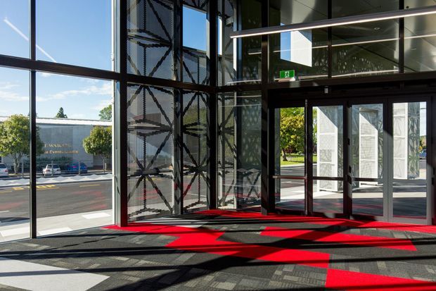 A bright, double-height glazed commercial entry lobby with expansive floor-to-ceiling windows, dark metal framing, automatic glass entrance doors, patterned glazing, and a bold red-and-black geometric carpet. Strong sunlight creates dramatic angular shadows across the floor, with an exterior street and landscaped grounds visible through the glazing.