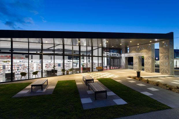 Contemporary commercial building viewed from a landscaped forecourt at dusk. The low, flat-roofed structure has extensive floor-to-ceiling glass curtain walls revealing a brightly lit retail interior, while a patterned perforated facade frames the entrance on the right. Grass panels, paved paths, outdoor benches, and integrated lighting define the public exterior space.