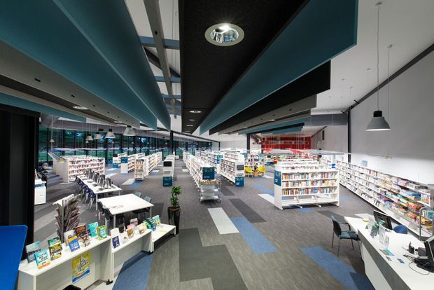 A large contemporary commercial retail interior, likely a bookstore or library, with long rows of white shelving filled with books and products. The space has a broad open floor plan, grey carpet tiles with blue and charcoal accents, white walls, extensive glazing along one side, suspended black pendant lights, and dramatic layered teal and black ceiling bulkheads. Tables, chairs, display stands, and service counters are distributed throughout the bright, organized space.