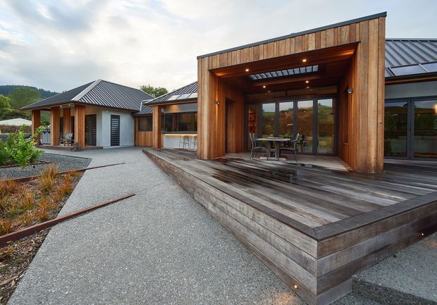 A single-storey contemporary home with a broad timber-clad entrance and covered outdoor entertaining deck. The building has dark metal pitched roofs, extensive glazing, warm timber columns and soffits, and a landscaped gravel forecourt with planted beds. A rural or bush-covered hillside is visible beyond the property.