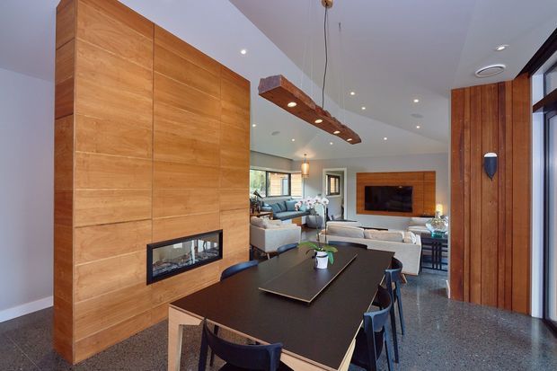 Open-plan contemporary interior combining a dining area and living room. A large timber-clad feature wall with a built-in fireplace separates the spaces, while a long dark dining table sits beneath a suspended timber-and-metal pendant. The living area includes a sectional sofa and a timber-framed media wall, with polished terrazzo-style flooring and extensive glazing.